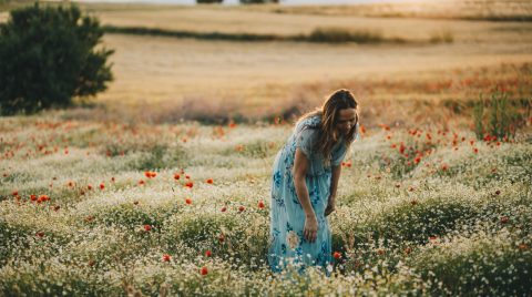 Mujer de pie, en un campo de amapolas con un vestido azul de flores, de espaldas al sol de atardecer.
